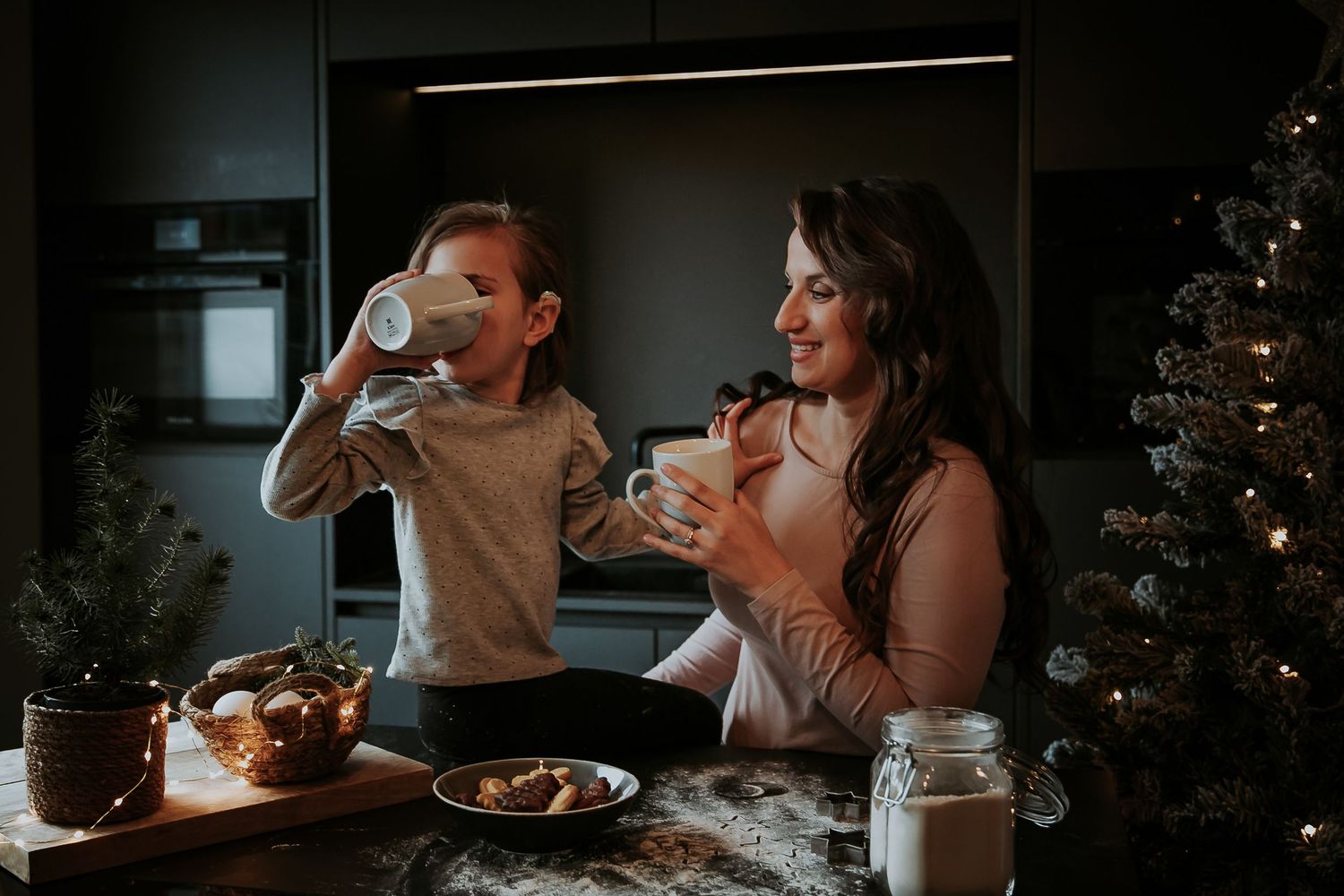 Smiling mother and daughter sip warm hot chocolate together in a cozy kitchen with a lit Christmas tree and fresh cookies on the counter.