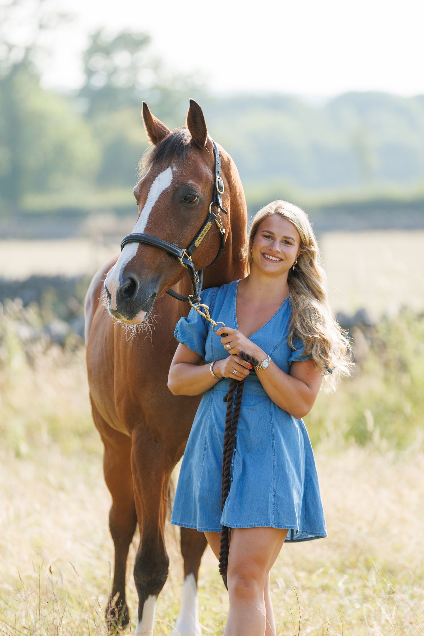 A man and woman smile at a farm gate surrounded by cattle and a horse in a lush green field.