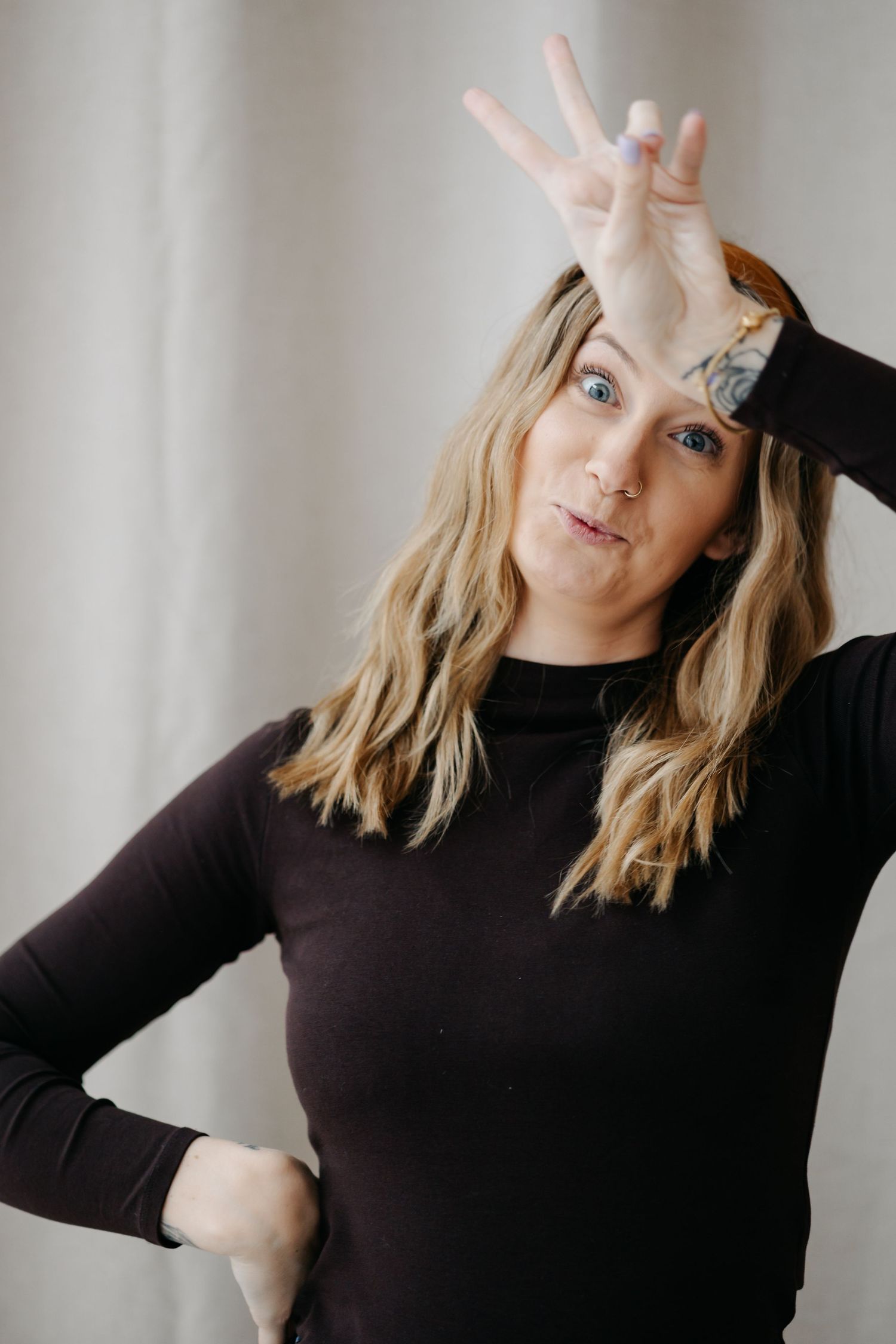 Woman in black turtleneck making silly face with hand gesture on forehead against light background.