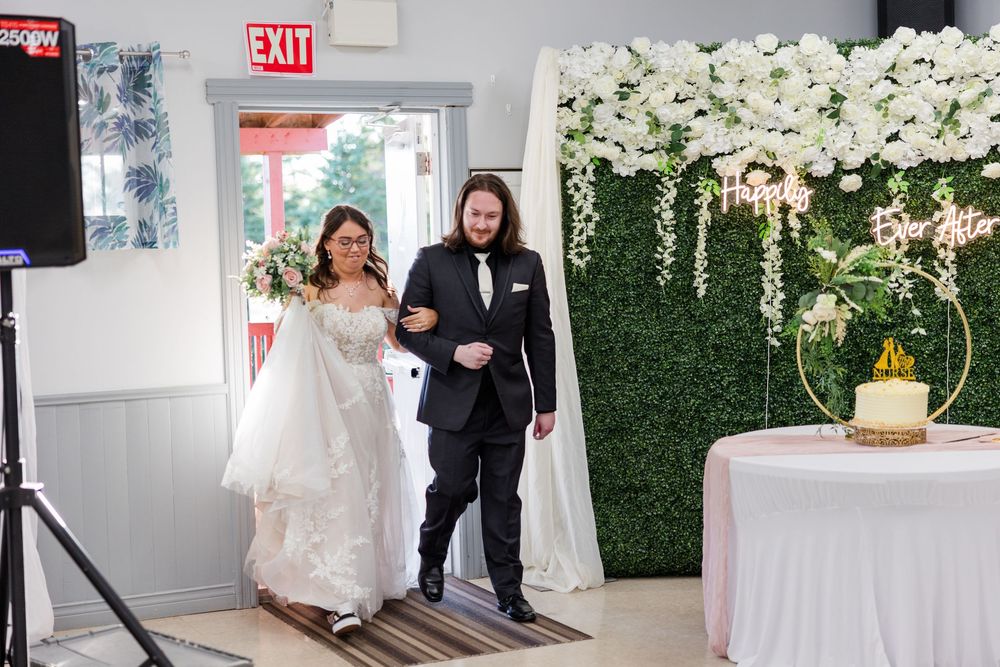 Bride and groom entering wedding reception venue with floral wall backdrop and white wedding cake on display.