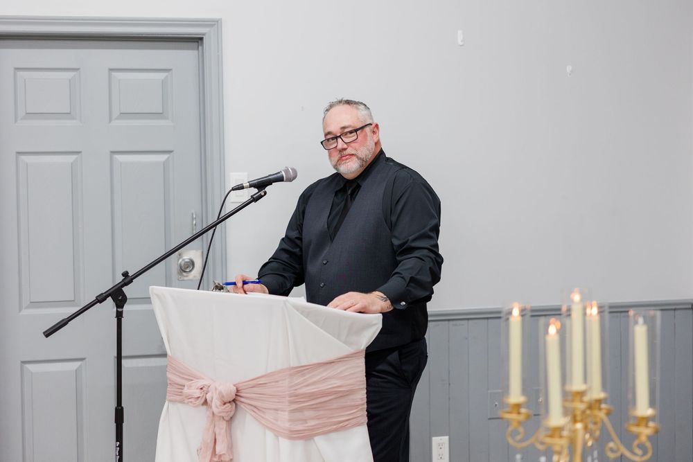 Man in black attire speaking at a decorated podium with microphone at a formal event.