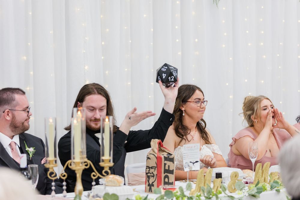 Wedding guest holds up a large black D20 dice at a reception table with candles and decorations.