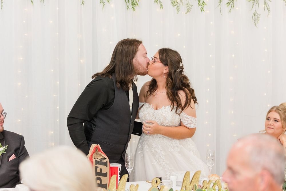 Bride and groom sharing a kiss at their wedding reception, with a white curtain backdrop and guests seated nearby.