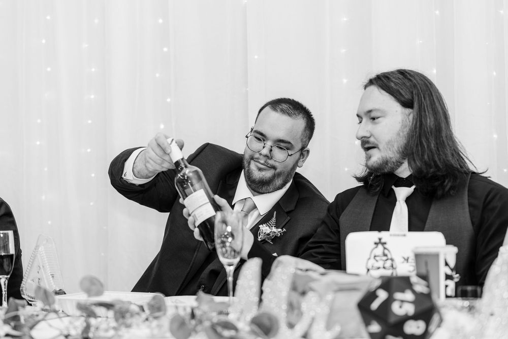 Two men at a wedding reception, one pouring a drink while the other reacts with surprise, captured in black and white.