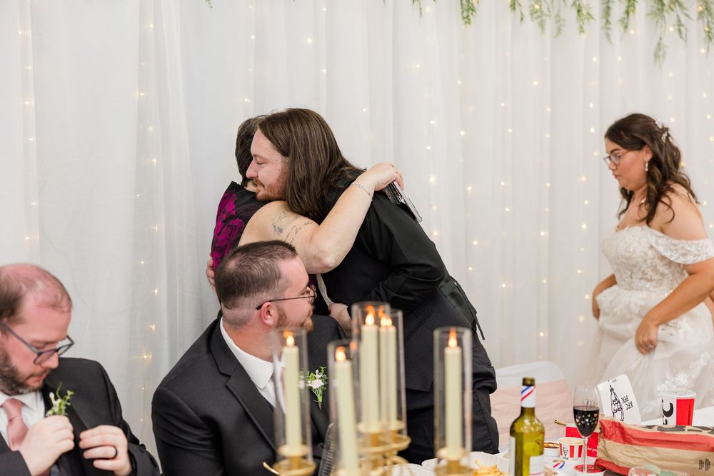Guests sharing a warm hug at a wedding reception, with the bride visible in a white gown in the background.