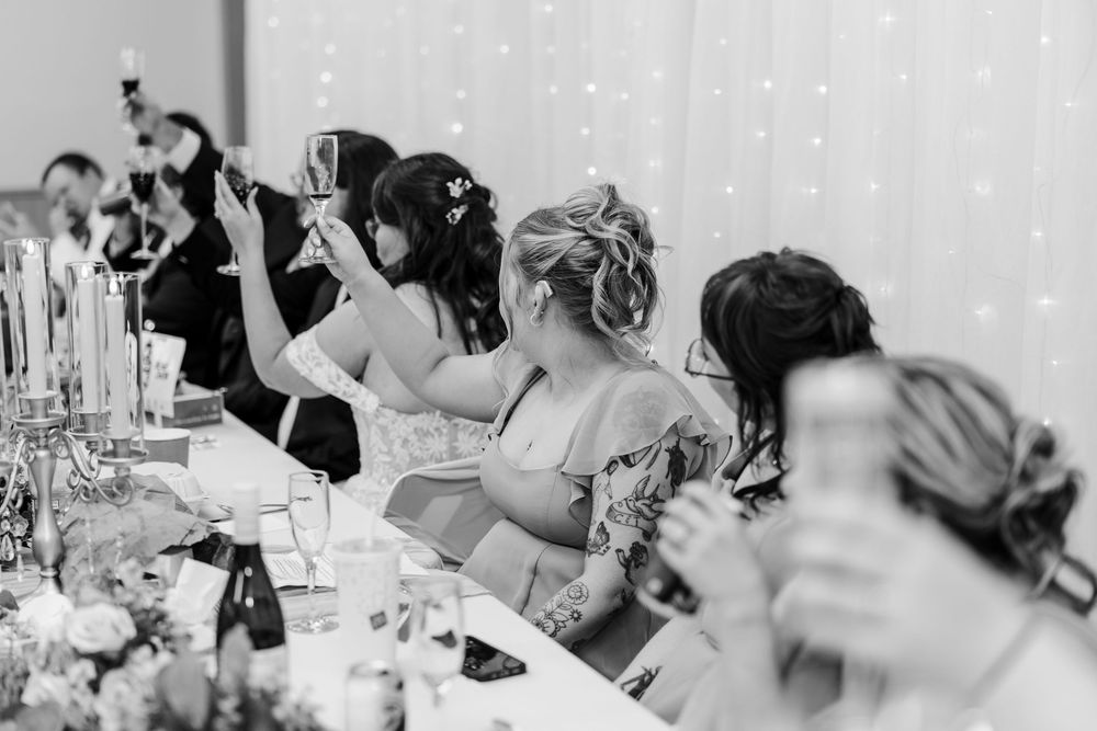 Black and white wedding reception photo of guests raising champagne glasses in a toast at an elegantly decorated table.
