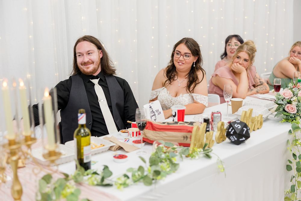 Wedding guests smiling at a decorated head table with candles, flowers, and bottles during a reception.