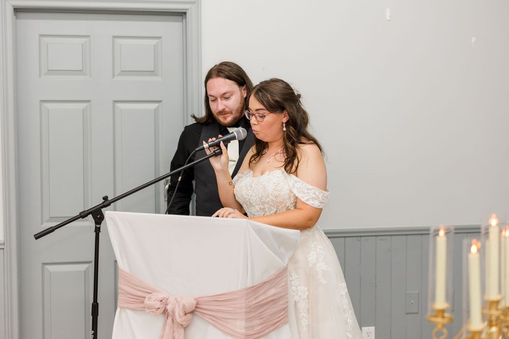 Bride and groom sharing a microphone at wedding reception podium, decorated with pink sash and candles.