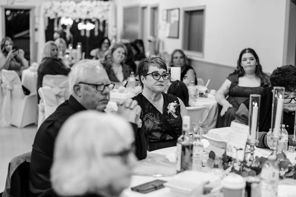 Black and white photo of wedding reception guests seated at tables, woman in glasses foreground.