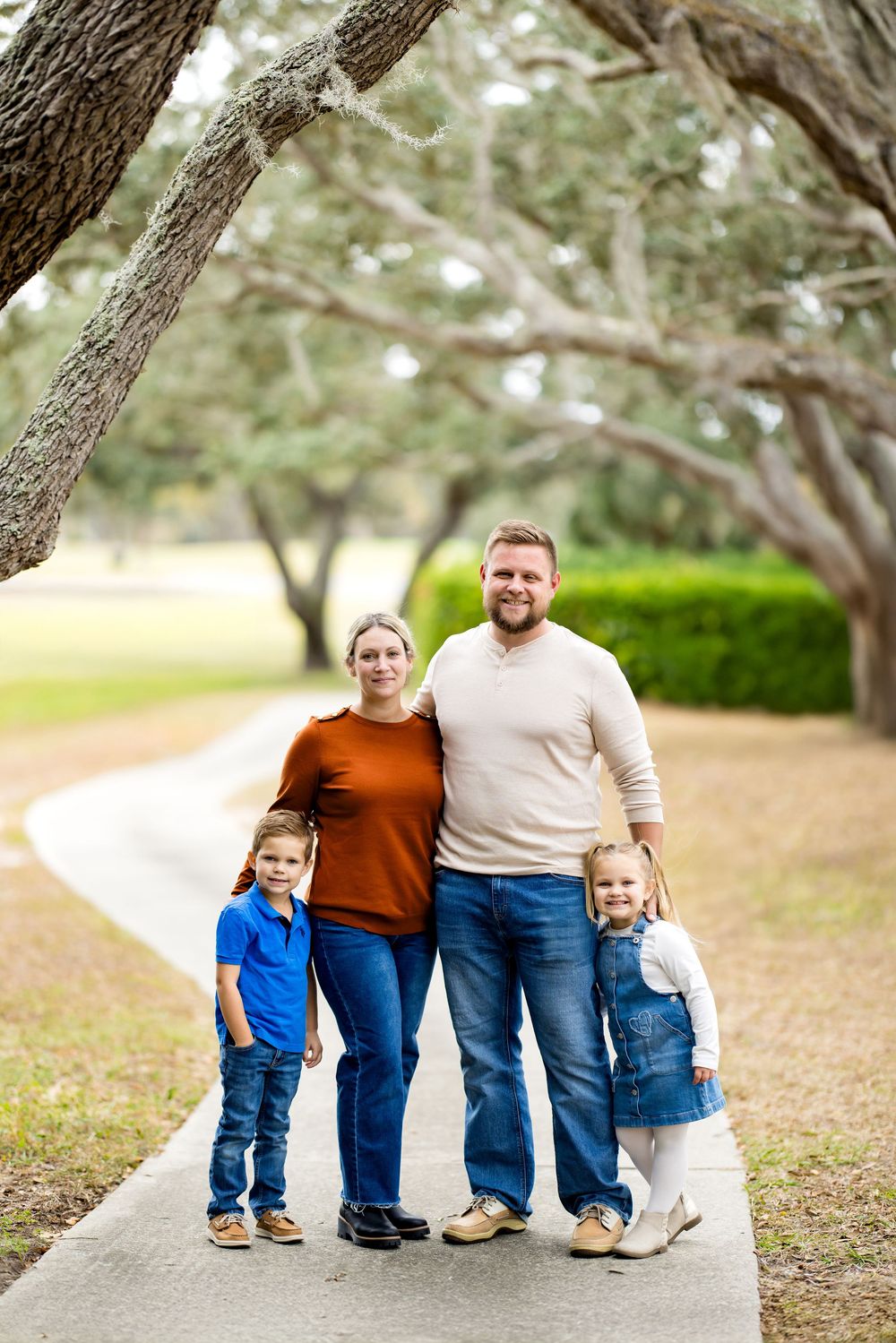 A smiling family of four poses on a tree-lined path in a park, wearing casual jeans and colorful tops.