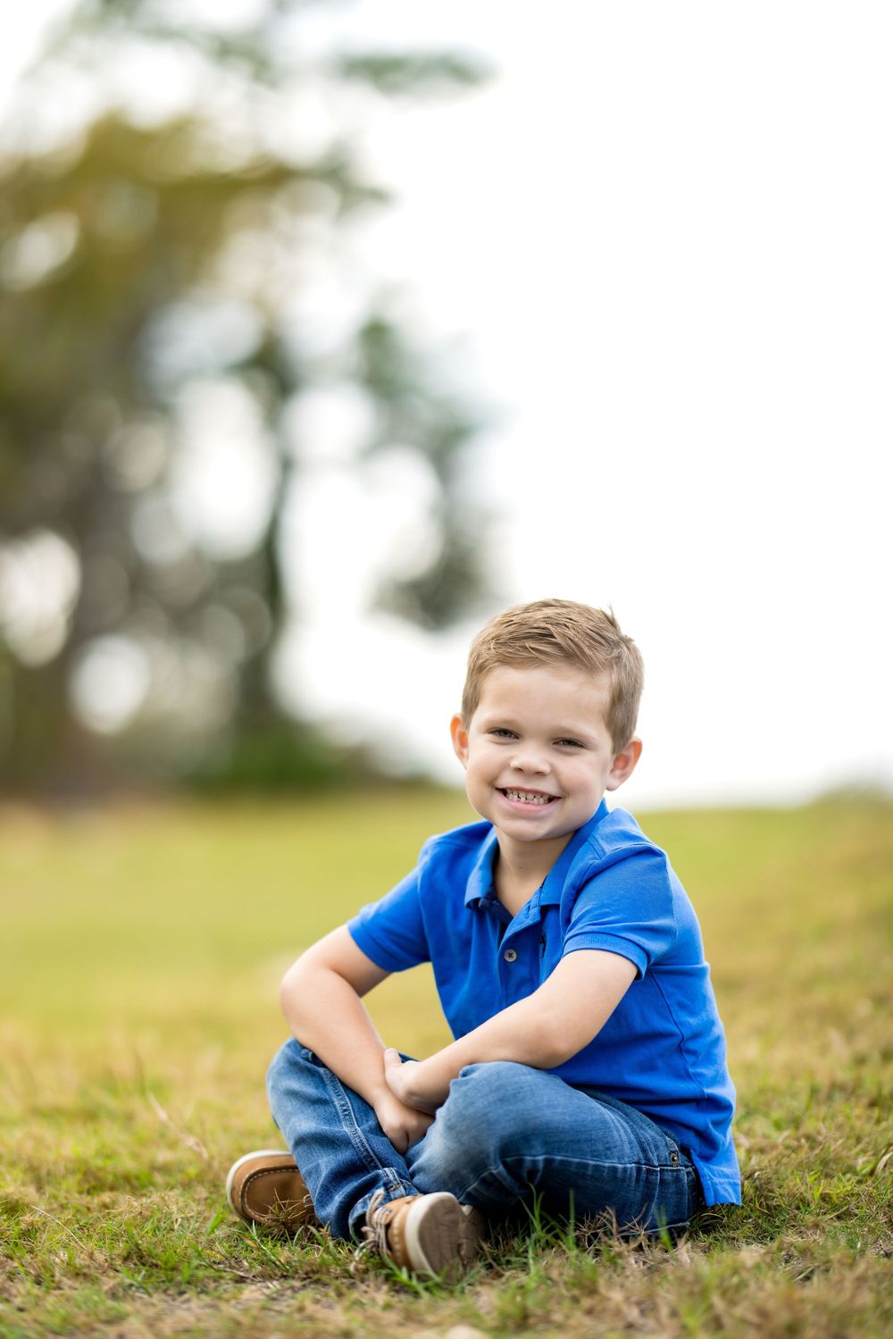 A smiling young boy in a blue polo shirt and jeans sits on green grass outdoors in a sunny park.