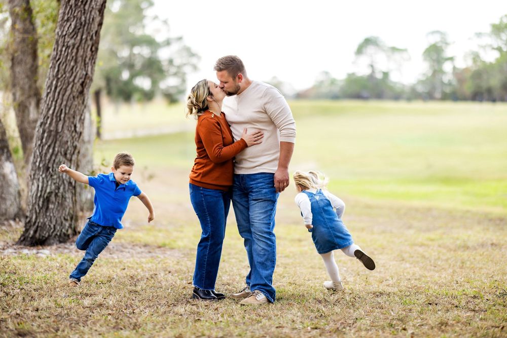 A happy family with two young children playing outdoors in a park, parents sharing a tender moment while kids run around them.