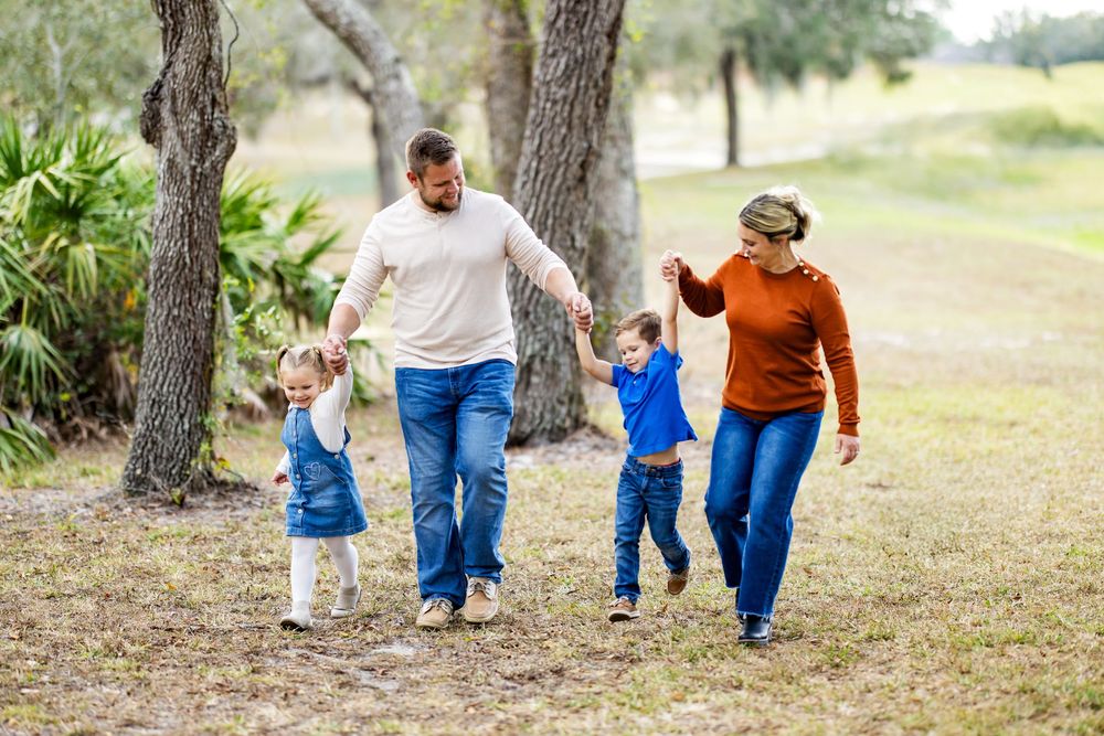 Family of four walking outdoors, parents swinging two young children by hands in a lush green park setting.