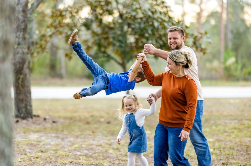Family playing outdoors in a park, parents swinging young boy in the air while toddler girl watches and laughs.