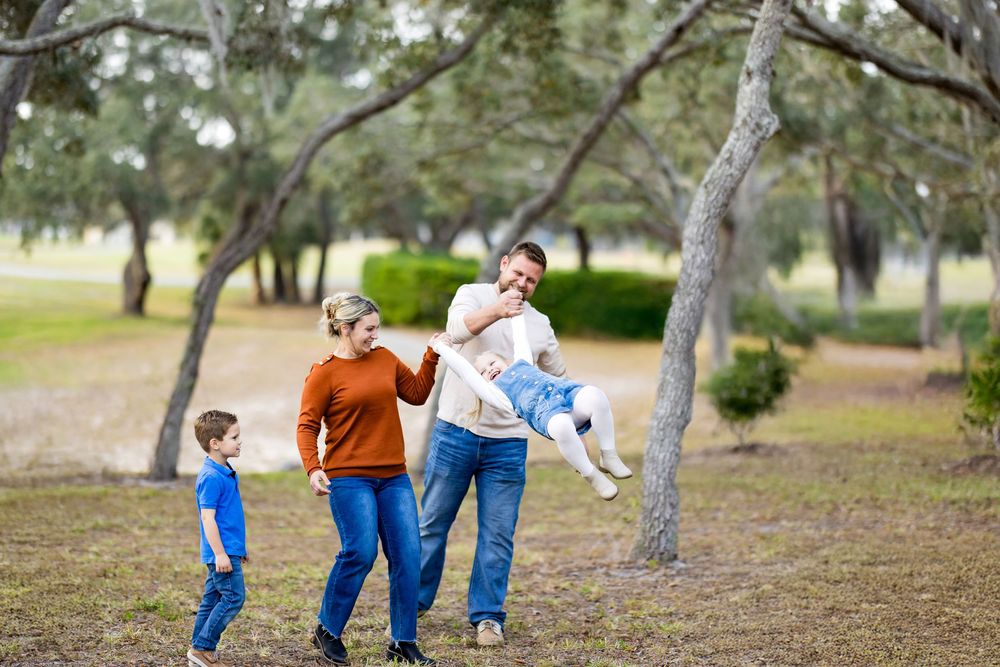 Family of four enjoying a sunny day outdoors in a park, parents playing with young children among trees.