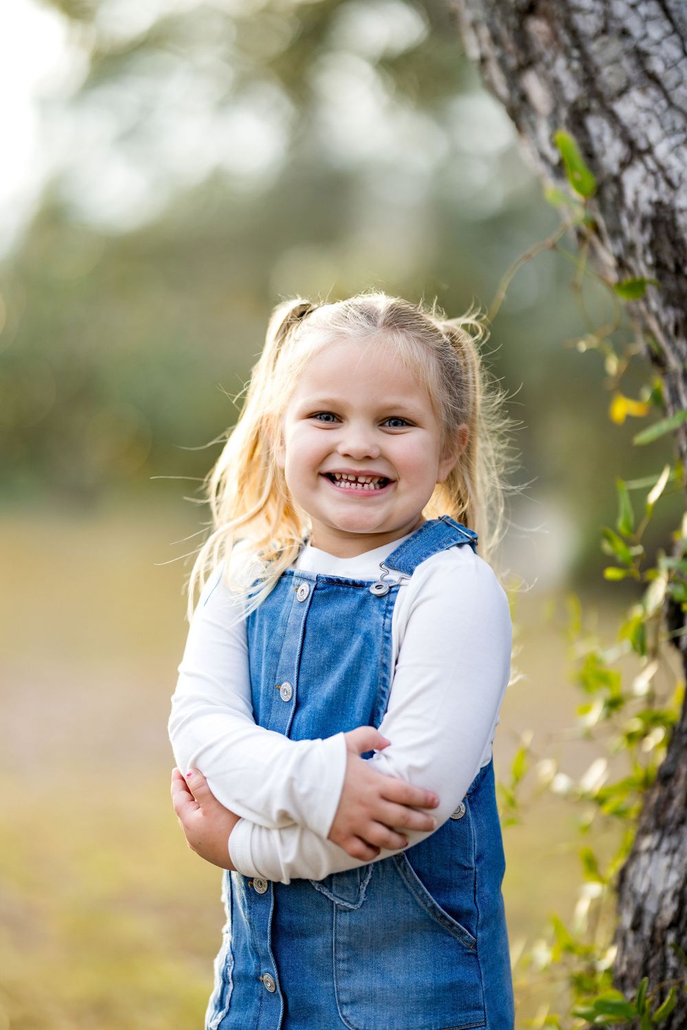 Smiling blonde toddler girl in denim overalls and white shirt stands outdoors near a tree with arms crossed.