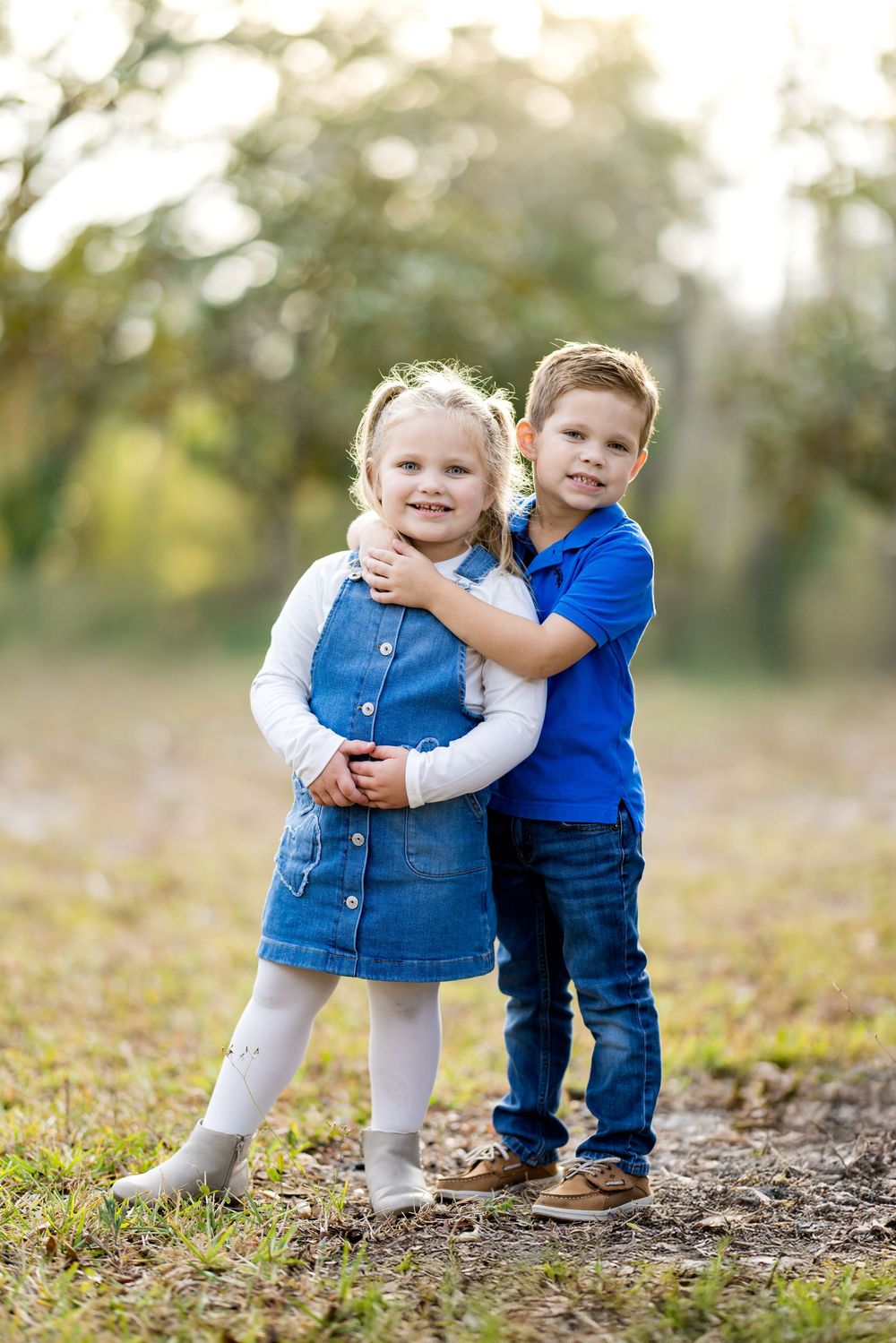 Two young children in denim outfits share a sweet hug outdoors, smiling in a sunlit green park setting.