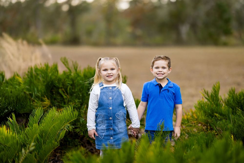 Two young siblings, a girl in denim overalls and a boy in blue polo, hold hands among green shrubs outdoors.