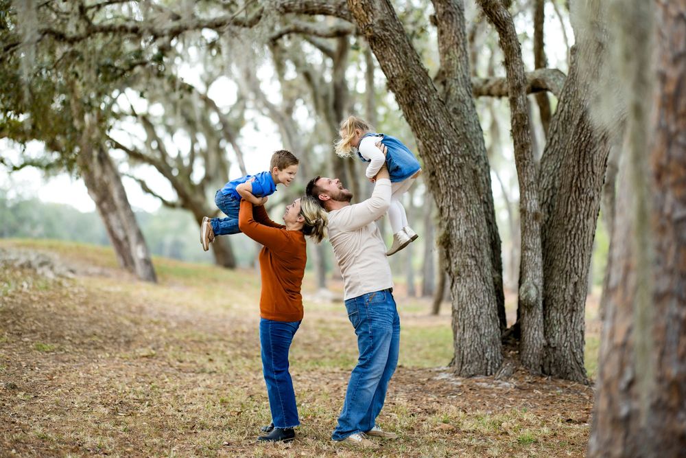 Two parents joyfully lift their young children in a scenic outdoor park surrounded by moss-draped oak trees.