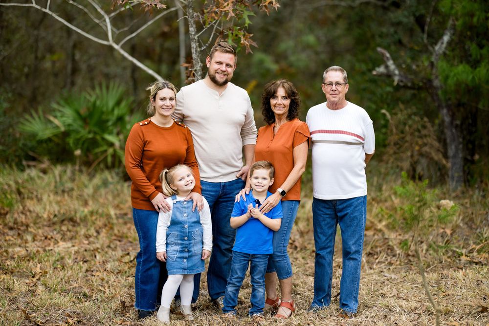 Multi-generational family of six posing outdoors in fall, wearing coordinating orange and blue outfits, with trees in background.