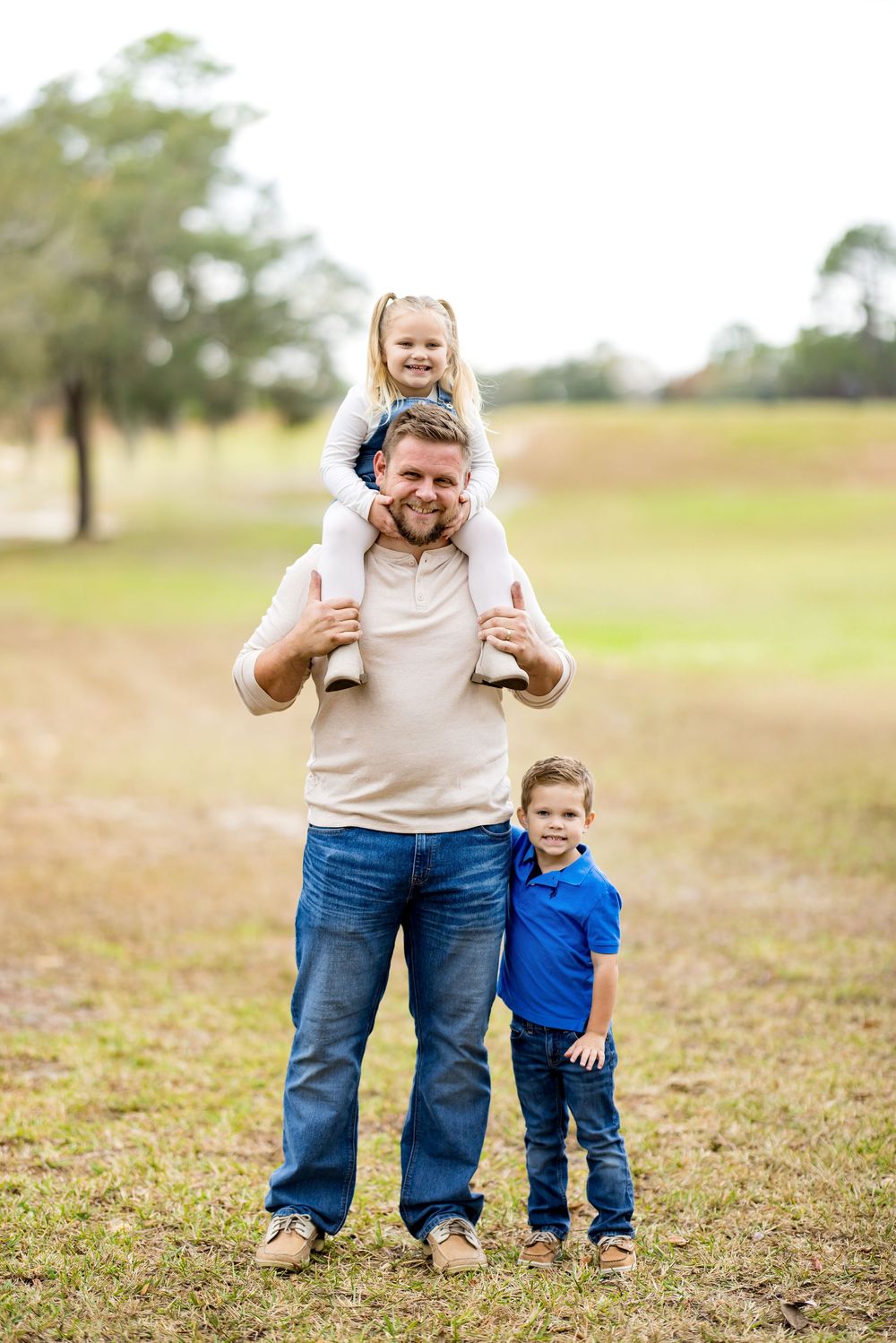A father smiles with his young daughter on his shoulders and son standing beside him outdoors in a grassy field.