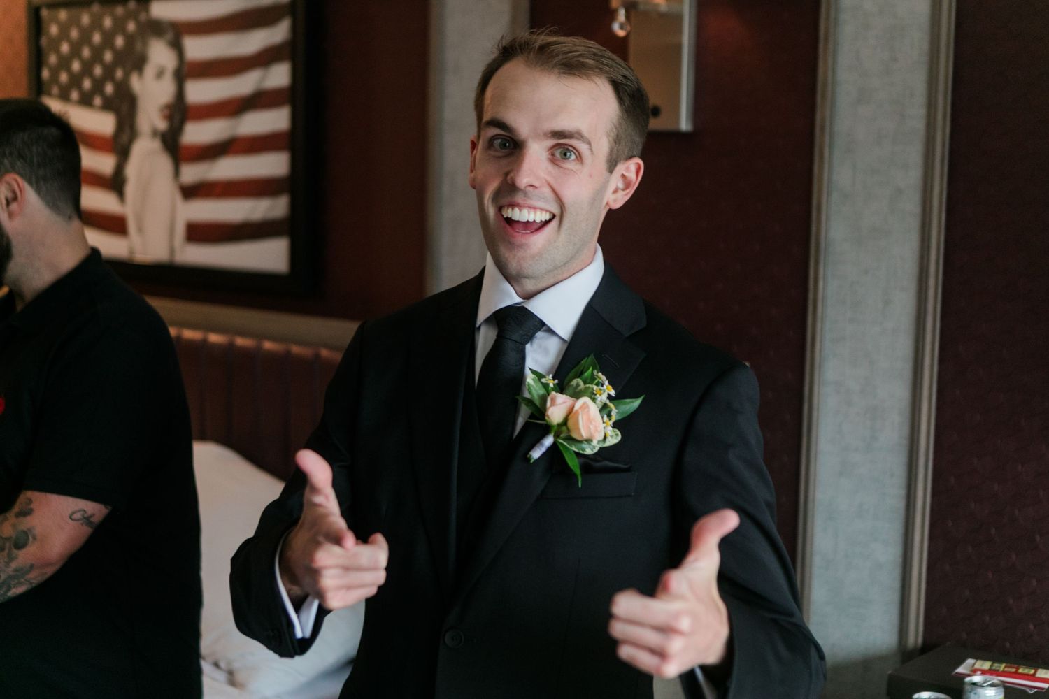 Smiling groom in black suit with boutonniere gesturing with open hands at wedding reception.