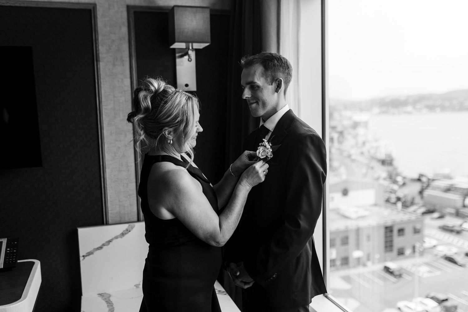 Black and white photo of a woman pinning a boutonniere on a man in a suit near a window with a city view.