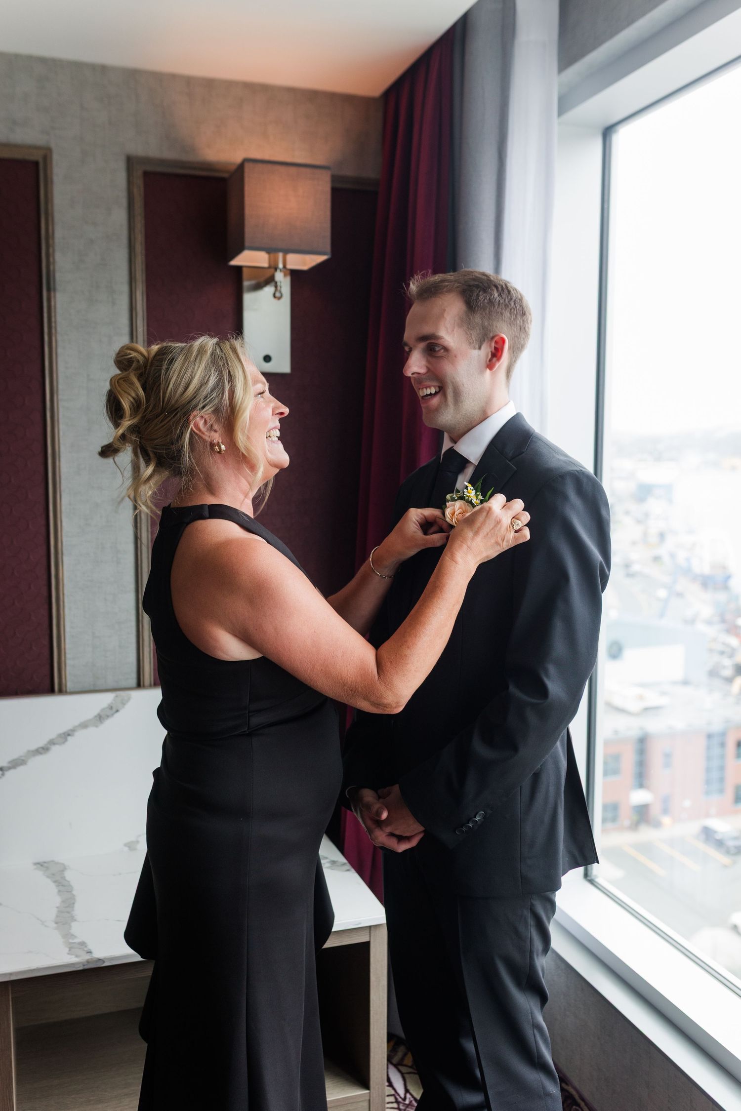 A woman in black dress pins a boutonniere on a smiling groom in a tuxedo near a bright hotel window.