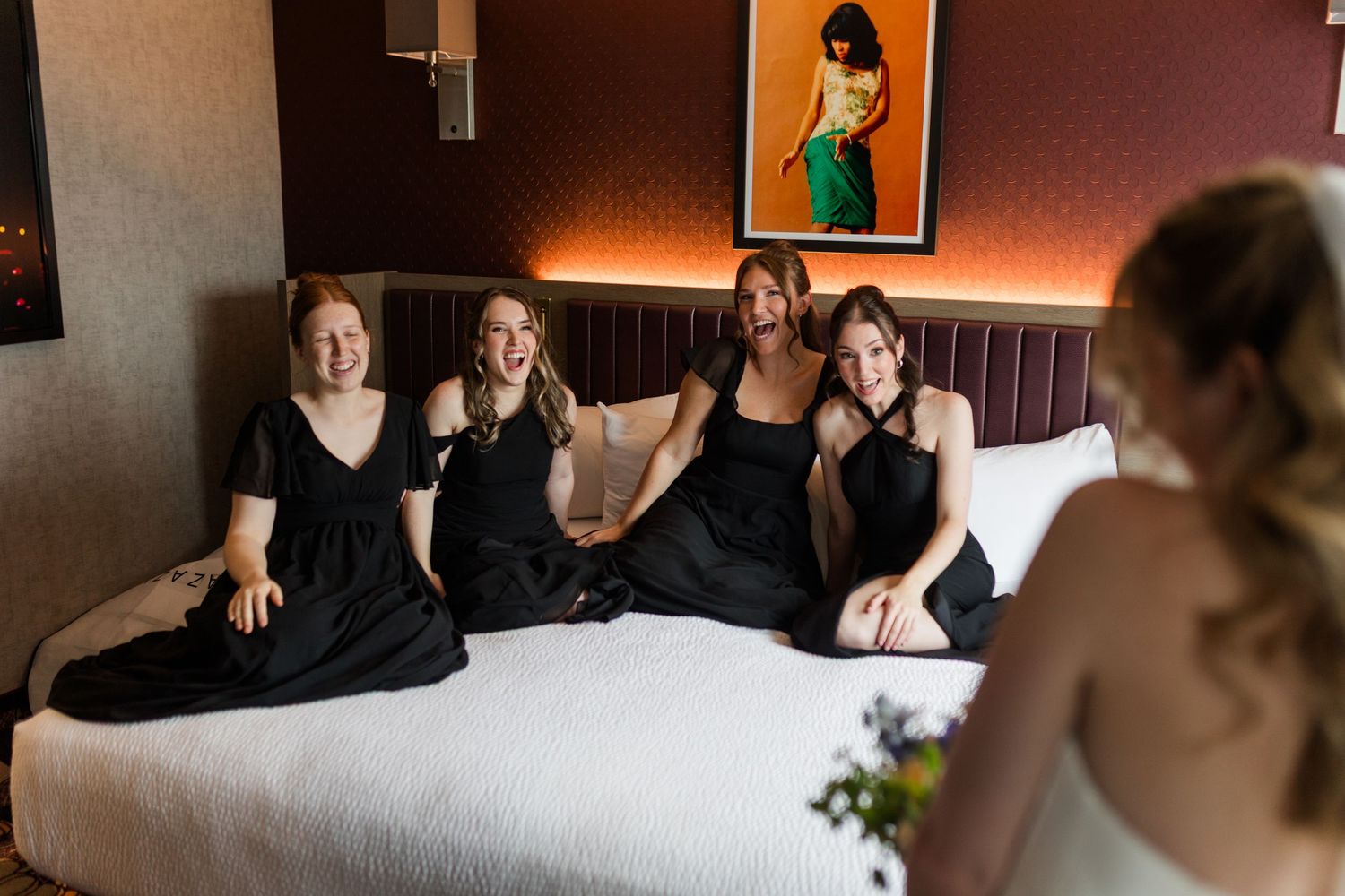 Four bridesmaids in black dresses sitting on a hotel bed smiling at a bride during wedding preparations.