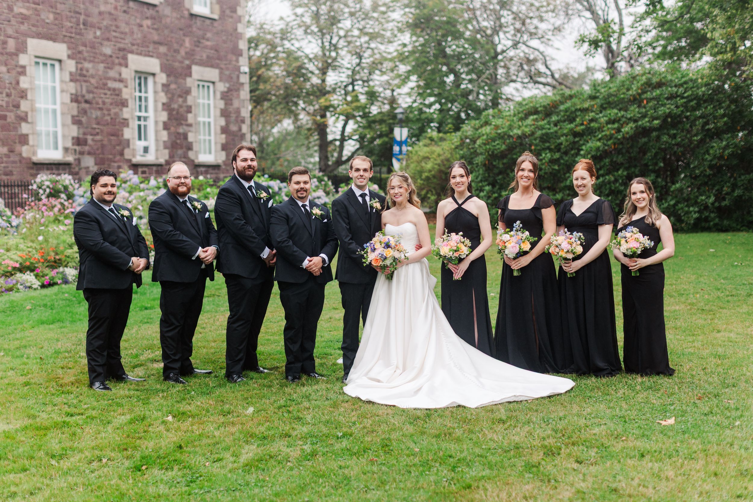 Wedding party in black attire poses with bride in white gown on green lawn outside stone building.