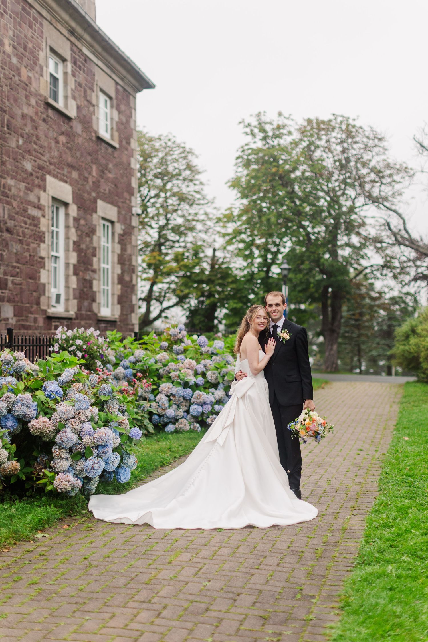 Bride and groom posing beside blooming hydrangeas near a historic stone building on their wedding day.