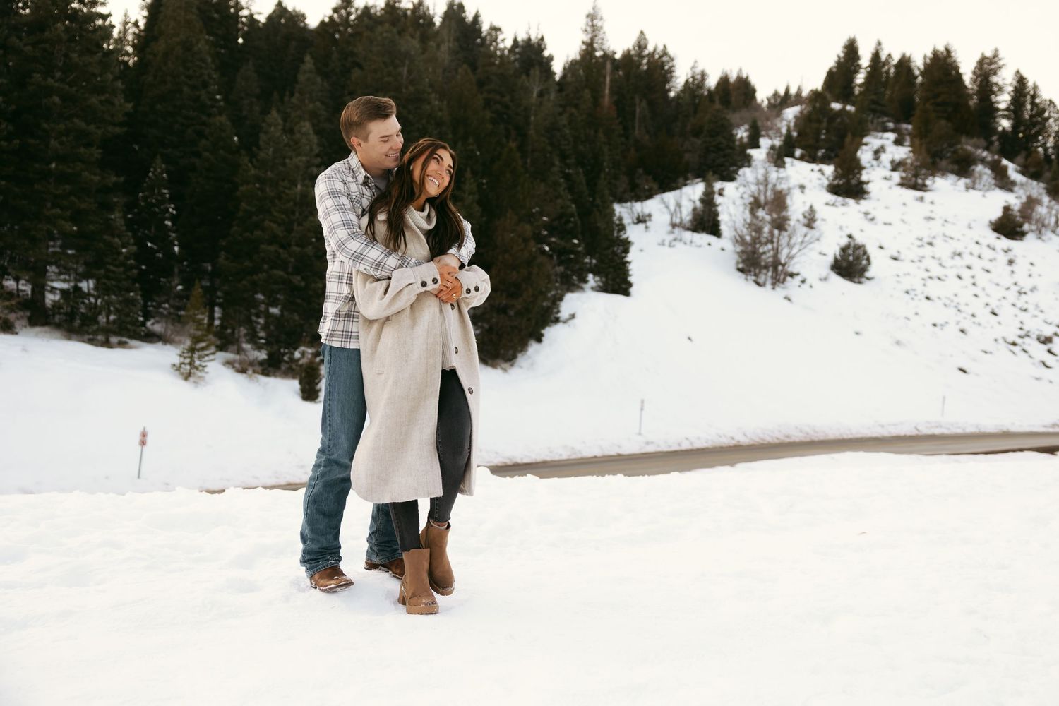 Man in Dusty Blue Flannel hugging woman in trench coat on winter snowy mountainside in warm clothes for engagement pictures
