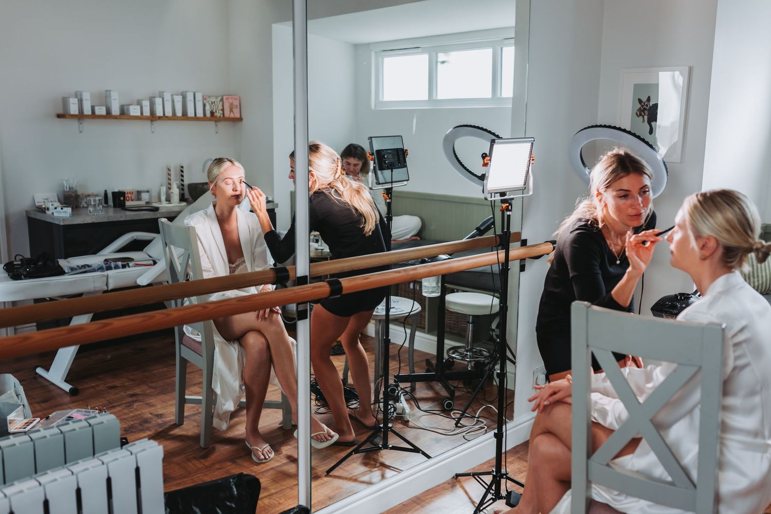 Women getting makeup done in a bright salon studio with ring light and mirror reflections.