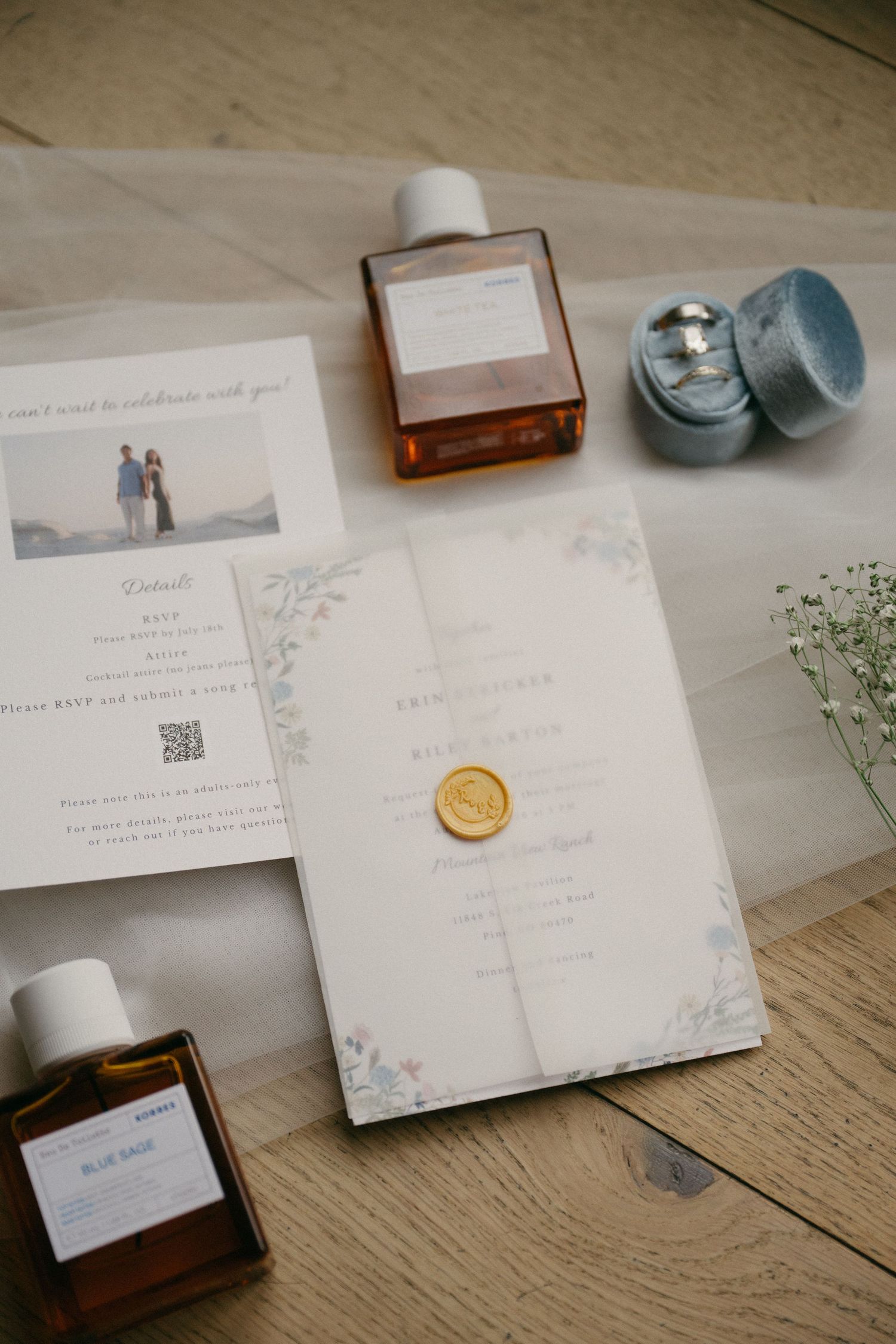 Wedding invitation suite with a gold wax seal, perfume bottles, and a velvet ring box on a wooden floor with tulle.