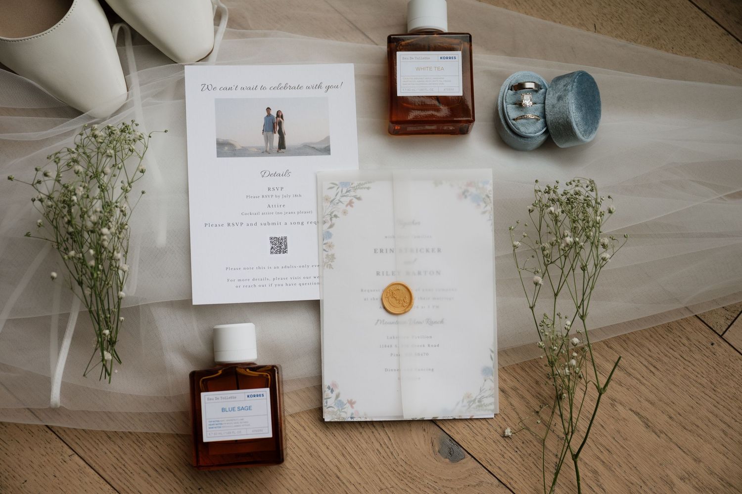 Flat lay of a vellum wedding invitation, amber perfume bottles, baby's breath, and a blue velvet ring box on tulle.