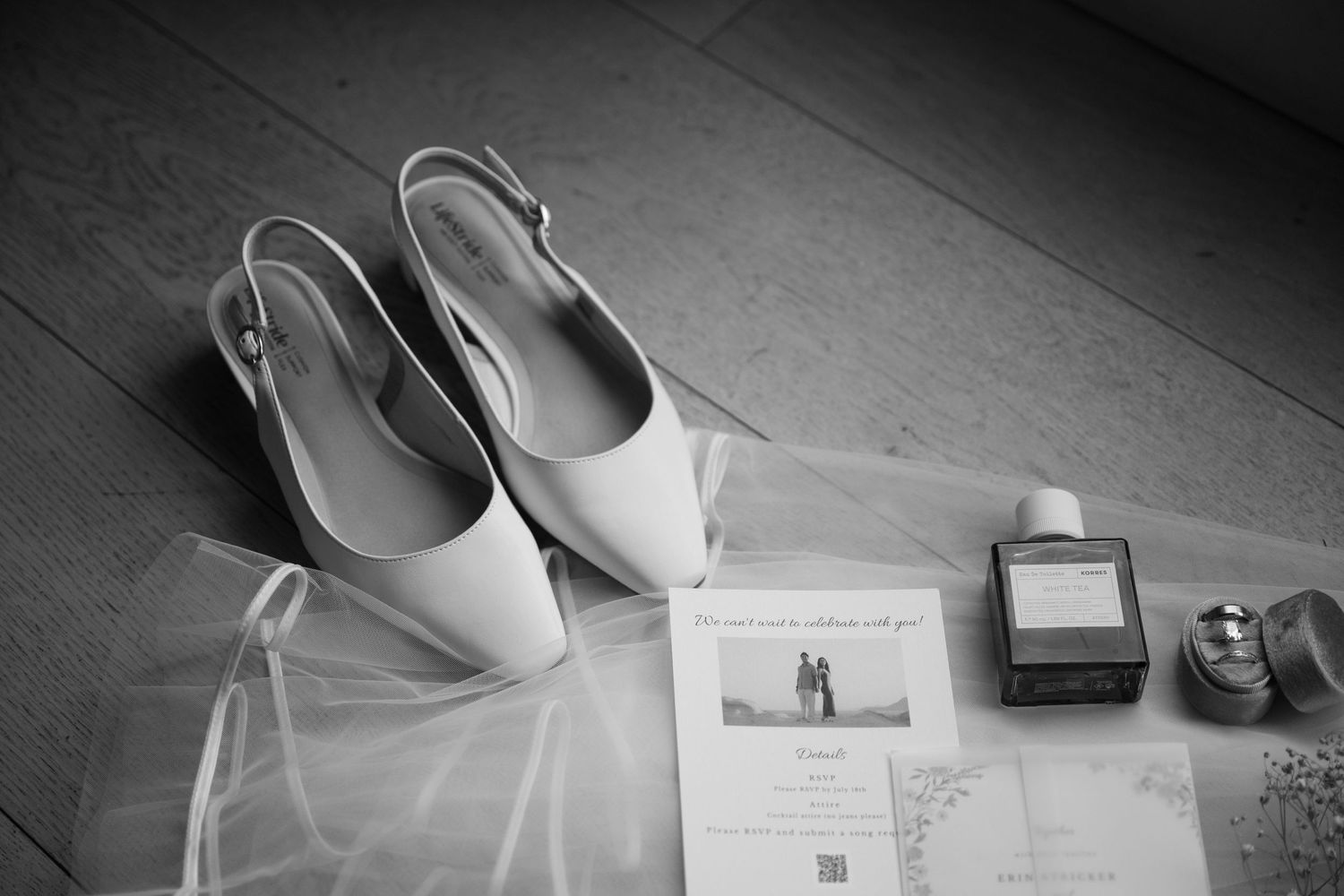 Black and white image of white bridal slingback heels beside a wedding invitation, perfume bottle, and ring box.