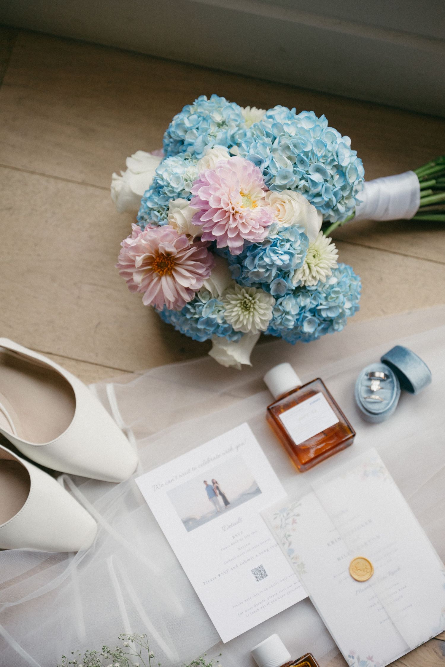 Blue hydrangea and pink dahlia bridal bouquet with white heels, perfume, ring box, and invitation on a wooden floor.