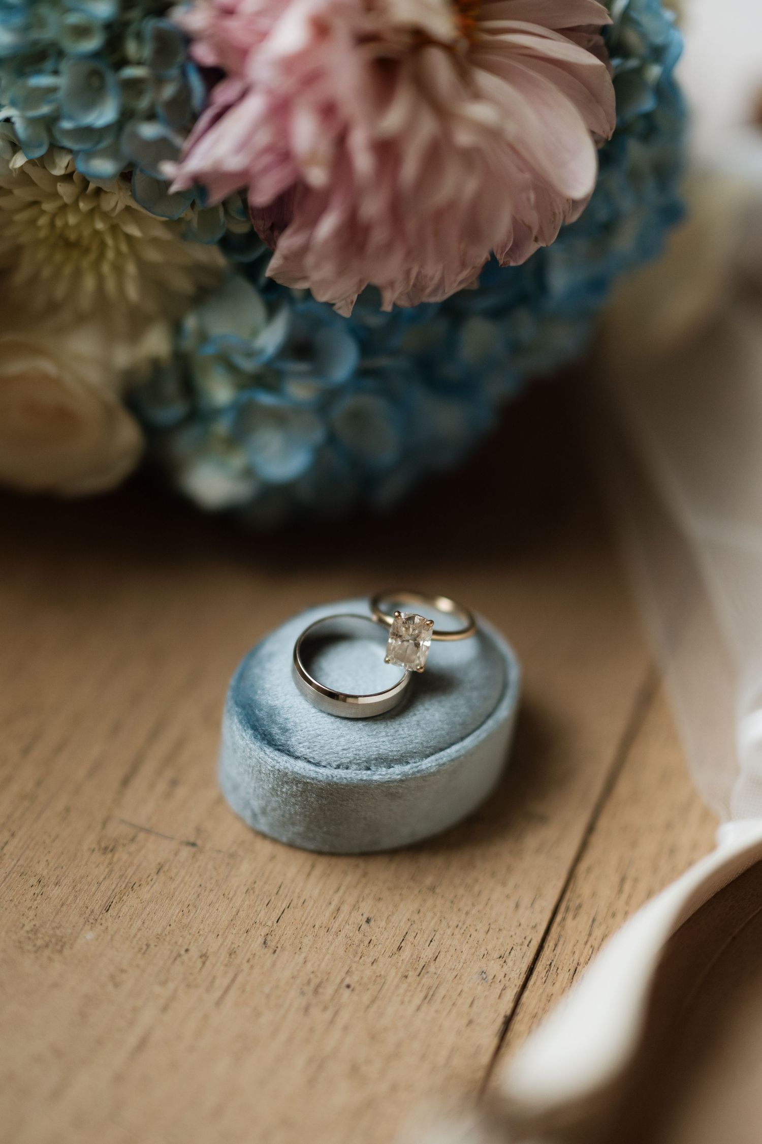 Two wedding rings, including a diamond engagement ring, rest in a blue velvet ring box near a blue and pink bouquet.