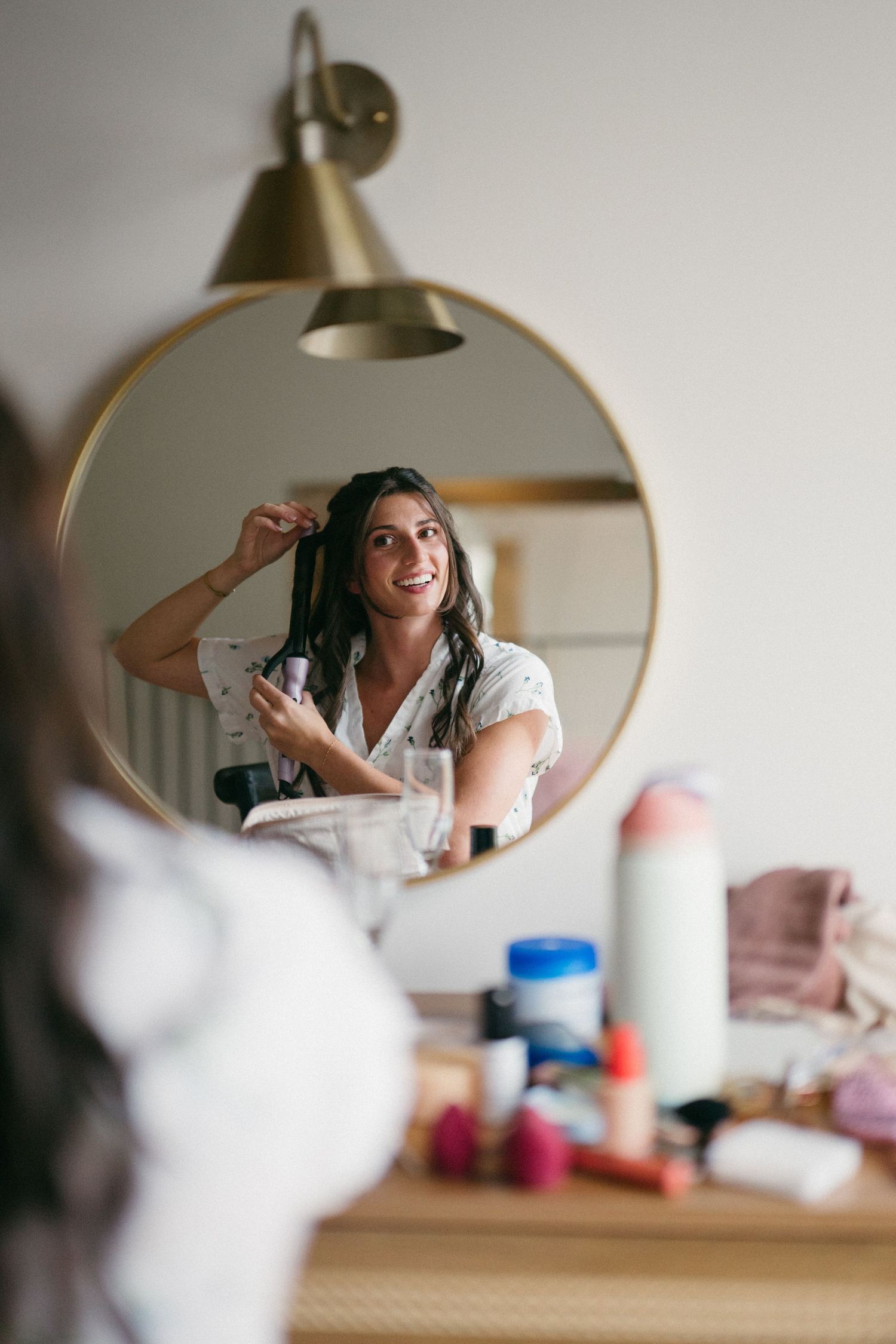 Smiling bride curls her hair with a curling iron while reflected in a round gold mirror on her wedding morning.
