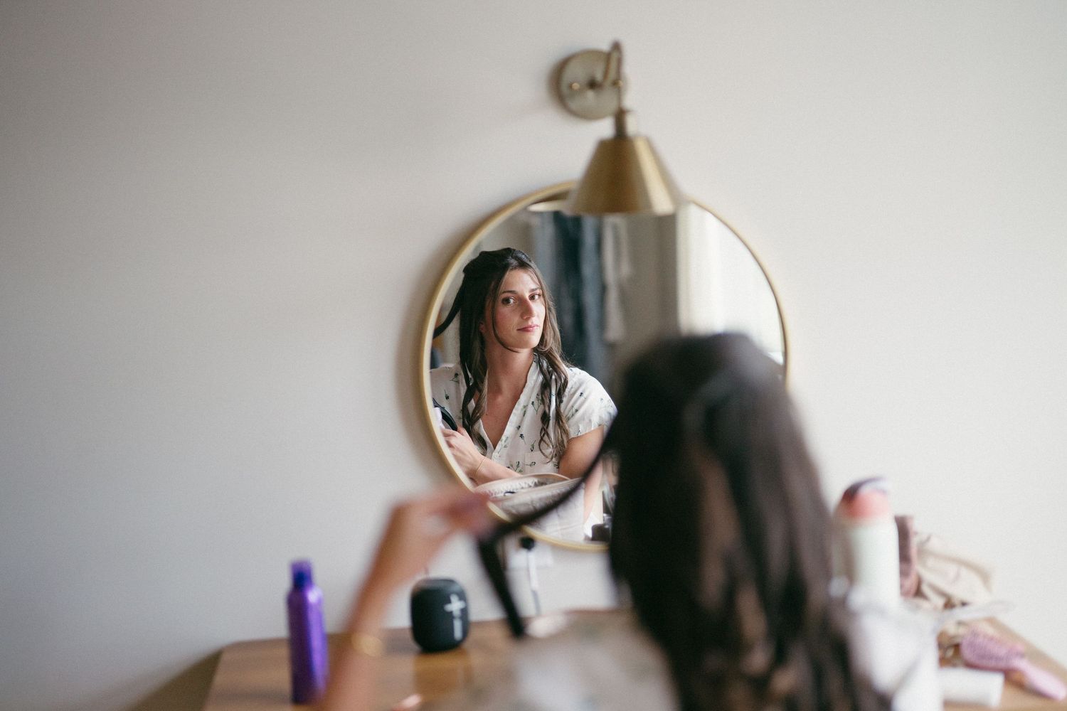Bride getting ready for her wedding looks into a round mirror while a hairstylist styles her dark hair.
