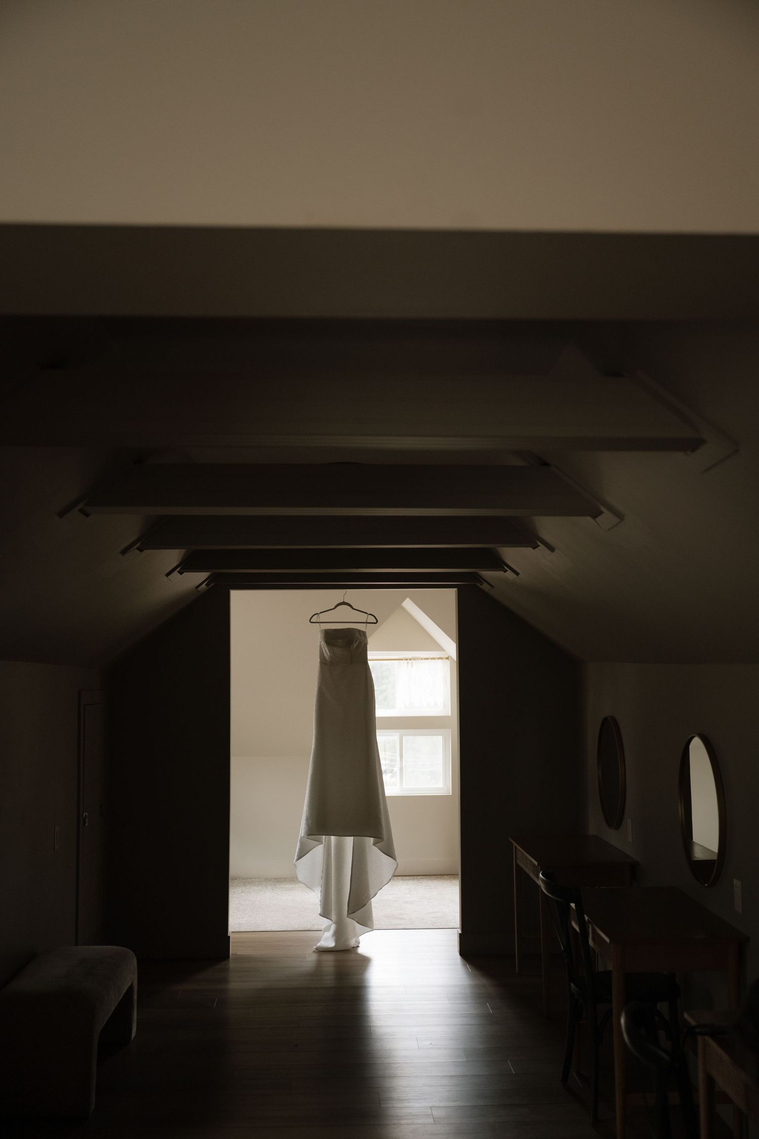 A white wedding dress hangs from a hanger in a doorway of a dimly lit room with exposed wooden beams and a bright window beyond.
