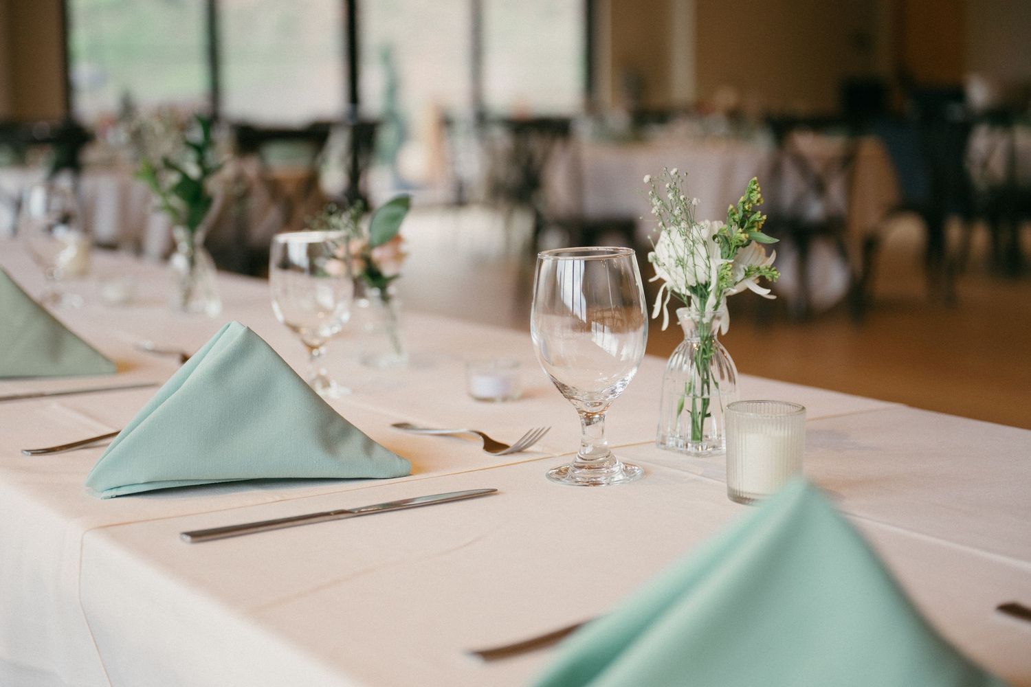 A wedding reception table set with sage green napkins, wine glasses, silverware, and small floral bouquets in the dining hall.
