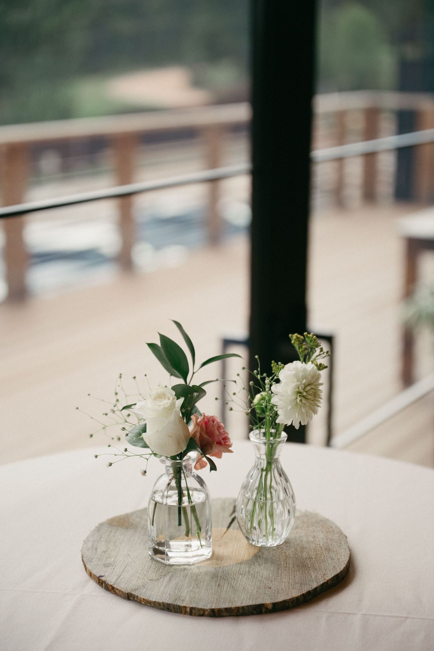 Two small glass bud vases with white and pink flowers sit on a wood slice atop a round wedding table beside a deck.