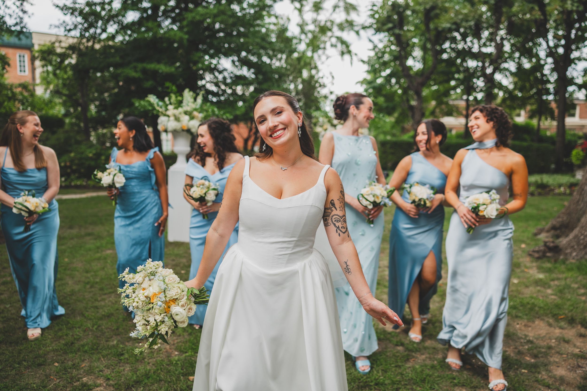 Bride in white gown smiles with bridesmaids in light blue dresses holding bouquets outdoors.