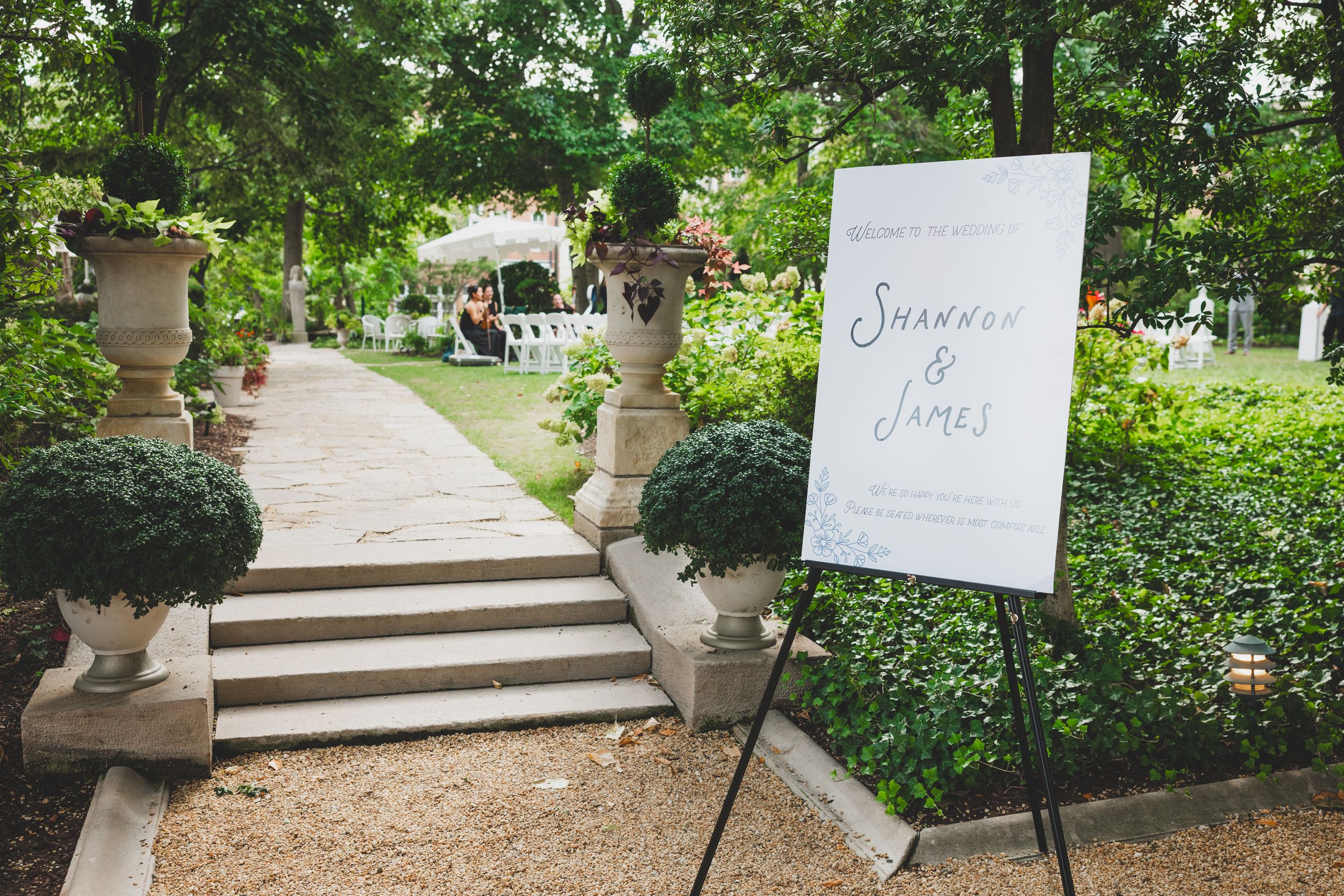 Outdoor wedding welcome sign for Shannon and James on an easel, surrounded by lush greenery and a stone pathway