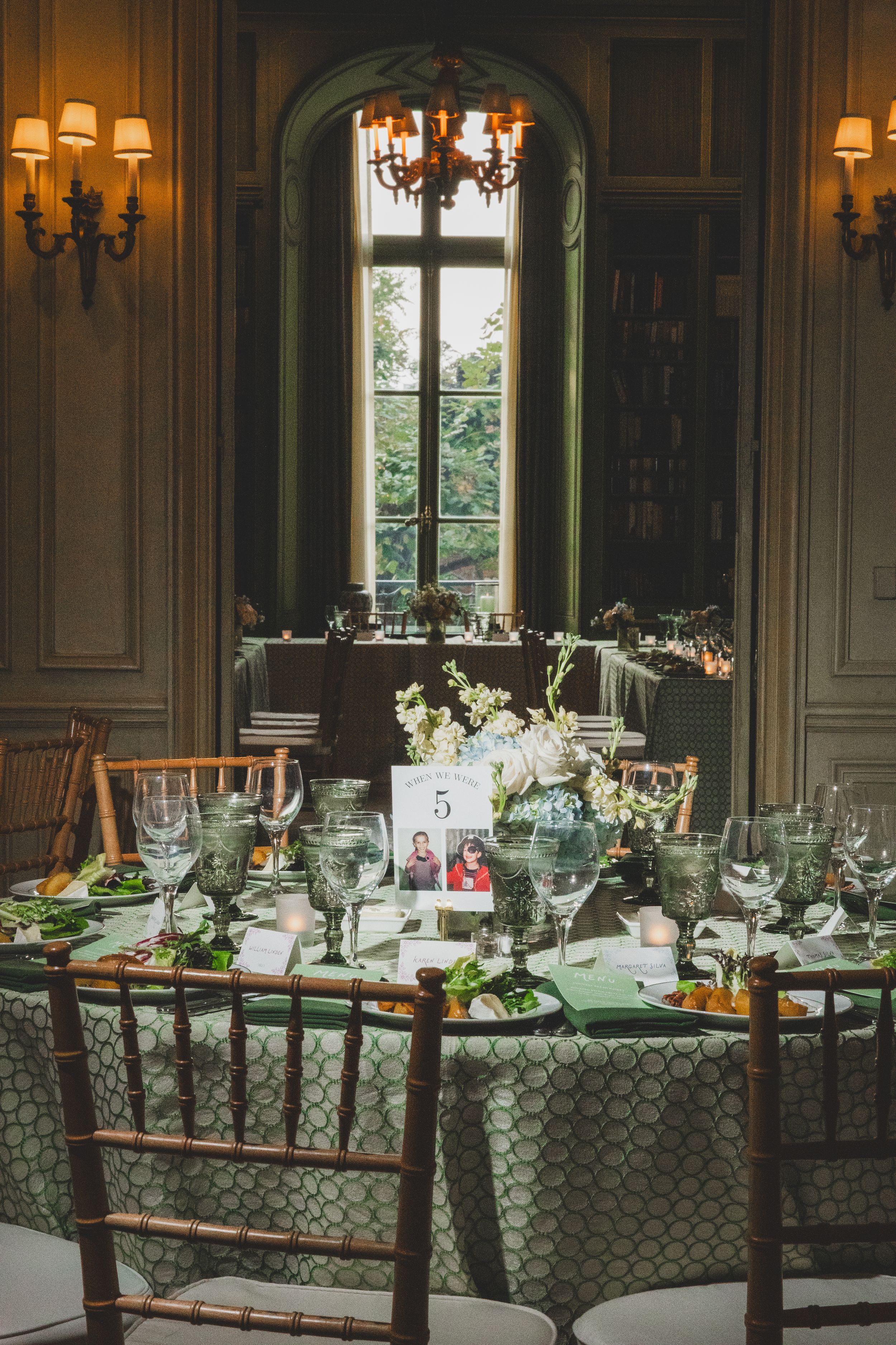 Elegant wedding reception table with green patterned linen, floral centerpiece, crystal glasses, and table number five in ornate room.