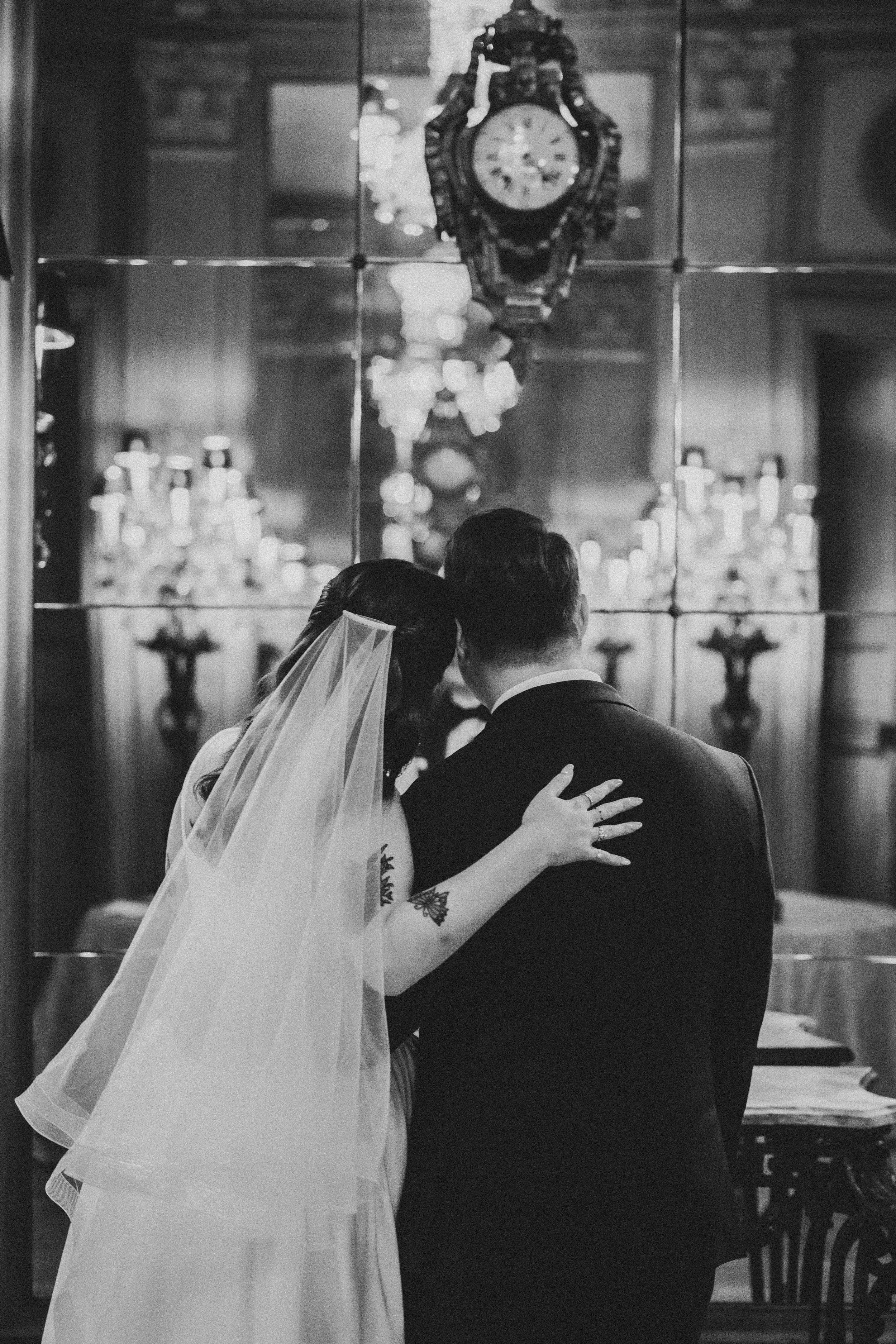 Black and white photo of a bride and groom embracing before an ornate clock and candelabras in an elegant venue.