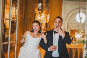Joyful newlyweds showing off wedding rings, bride in white gown holding cocktail, groom in tuxedo, elegant venue.