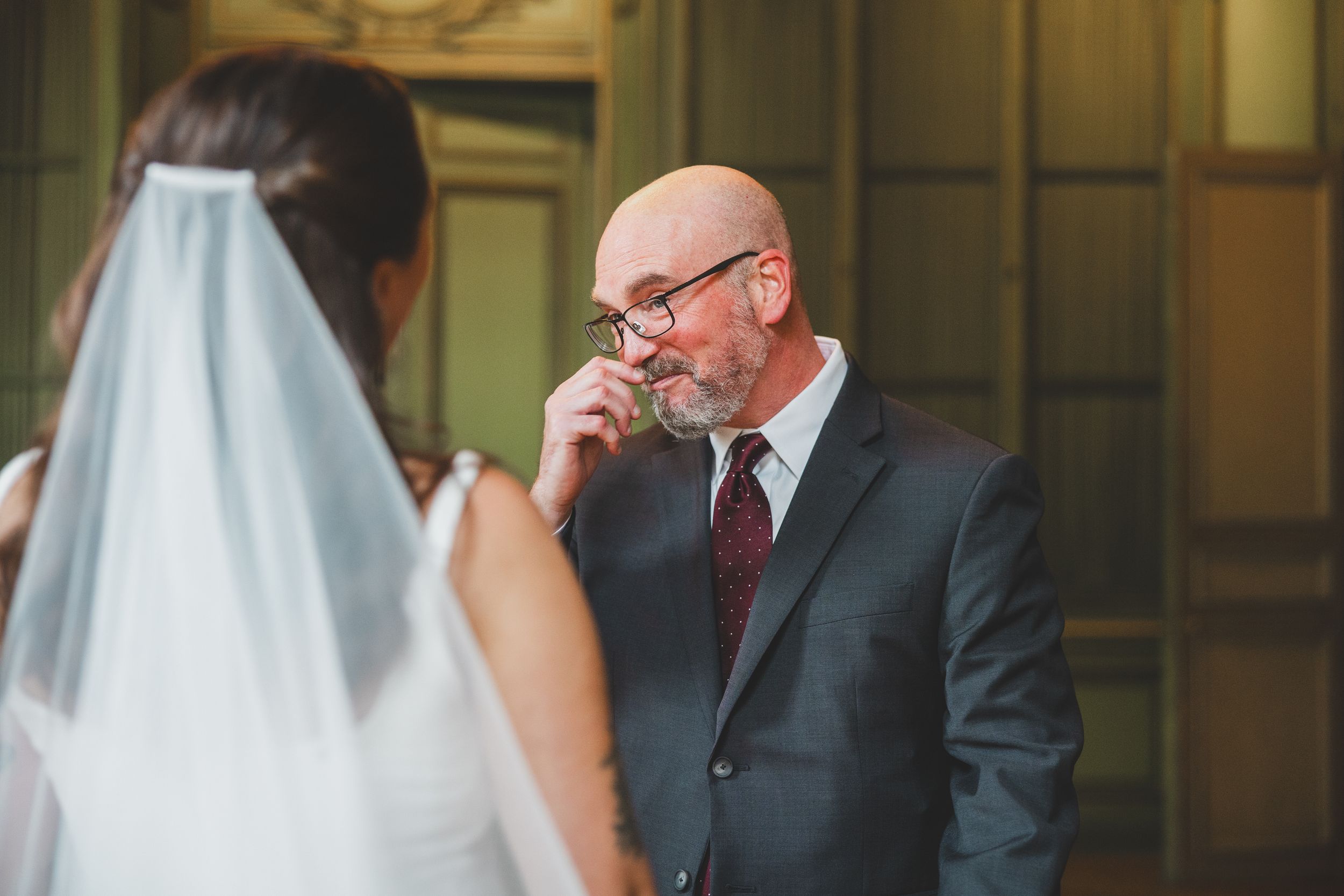 Emotional bald man in gray suit wiping tear at wedding ceremony, seen from behind bride in white veil.