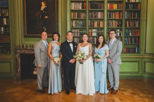 Wedding party poses in elegant library with green walls, bookshelves, bride in white gown, bridesmaids in blue dresses.